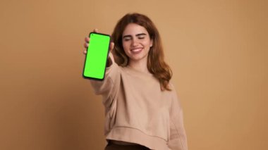 Siyah saçlı, günlük kıyafetli mutlu bir kadın kameraya bakarken yeşil ekranlı akıllı telefonu bej arka planda duruyor.