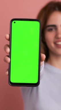 Siyah saçlı, rahat giyimli mutlu bir kadın kameraya bakarken pembe arka planda duran yeşil ekranlı akıllı telefonu gösteriyor. Dikey Görüntü