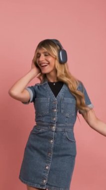 Kot giymiş neşeli transseksüel kadın pembe arka planda müzik dinlerken kameraya bakıyor.