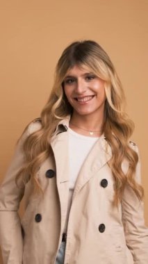 Trençkotlu mutlu transseksüel kadın bej arka planda dururken kameraya bakıyor.