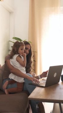 Hafta sonu rahat bir oturma odasında kanepede çizgi film izlerken küçük kızına bilgisayarı gösteren gündelik kıyafetlerle gülümseyen Latin anne.