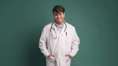 Gülümseyen genç İspanyol erkek konuşuyor ve kameraya bakıyor. Üzerinde steteskop ve elleri cebinde yeşil arka planda dururken.