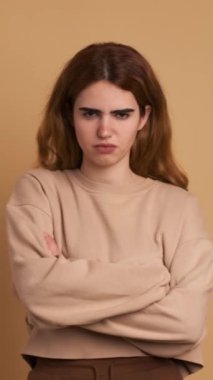 Kollarını kavuşturup kameraya bakan kızgın bir genç kadının kahverengi arka planlı stüdyo videosu. Dikey Görüntü