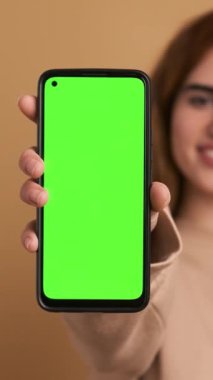 Gayri resmi giyinmiş, siyah saçlı mutlu bir kadın kameraya bakarken yeşil ekranlı akıllı telefonu bej arka planda gösteriyor. Dikey Görüntü