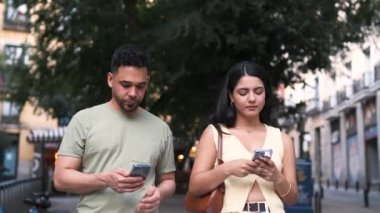 Gündüz vakti sokakta yürürken akıllı telefonlar kullanan, günlük giysiler içinde sıkılmış, genç İspanyol bir çift.