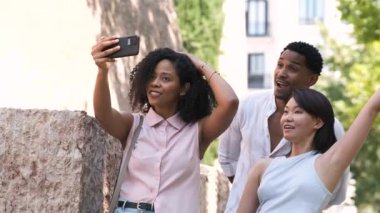 Neşeli, çokkültürlü erkek ve kadın arkadaşlar, güneşli bir günde şehirde akıllı telefondan selfie çekerken rahat kıyafetler giyip dillerini çıkarıyorlar.