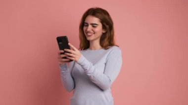 Aynı anda hem gülümseyen hem de cep telefonu kullanan mutlu bir kadının pembe arka planına sahip bir stüdyo videosu.