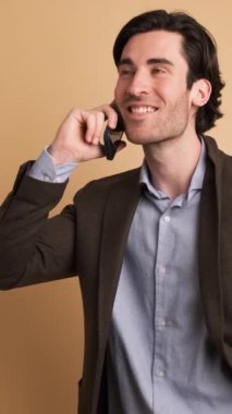 Siyah saçlı, gülümseyen, cep telefonuyla konuşan ve kahverengi arka planda kameraya bakan yetişkin iş adamının portresi.