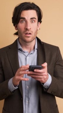 Takım elbiseli gülümseyen işadamının portresi, kahverengi arka planda dururken akıllı telefondan iyi haberler aldıktan sonra şaşırmış ve kutlama yapıyordu.