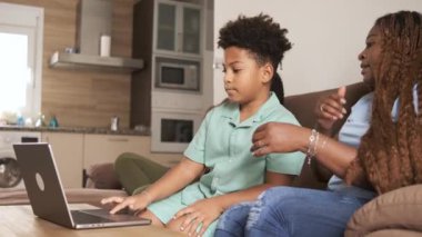 Anne, evdeki oturma odasında dizüstü bilgisayar kullanırken oğluna sevgiyle yardım ediyor. Sıcak bir ortamda birlikte otururken ona sabır ve özenle rehberlik ediyor.