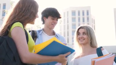 Güneşli bir günde üniversite kampüsünde ayakta durup kitap okuyarak bir projeyi tartışan günlük giysiler içinde gülümseyen genç erkek ve bayan öğrencilerin ağır çekimde