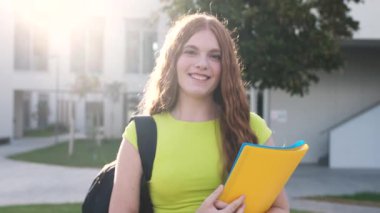 Güneşli yaz gününde üniversite kampüsünde kameraya bakarken uzun kızıl saçlı gülümseyen genç kız öğrencinin portresi.