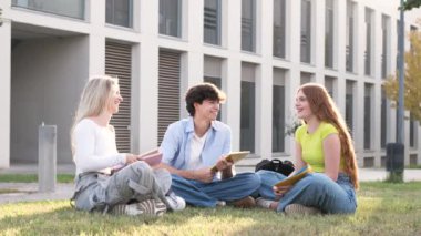 Güneşli bir günde kampüste çim üzerinde kitap ve tabletle oturmuş sohbet eden neşeli erkek ve kadın öğrencilerin karantinaya alınmış fotoğrafları.