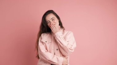 Pembe kot ceketli, uzun kahverengi saçlı, öpüşen ve pembe arka plandaki kameraya bakarken kalp hareketi yapan mutlu genç bir kadının portresi.