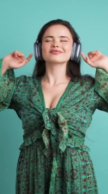 Kahverengi saçlı, yeşil elbiseli mutlu genç bayan mavi arka planda kablosuz kulaklıklarla dans müziği dinliyor ve eğleniyor.