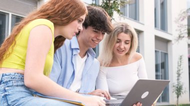 Bir grup öğrenci dışarıda oturuyor, bir dizüstü bilgisayarda birlikte çalışıyorlar. Fikirlerini paylaştıkları ve ekrana odaklandıkları için, işbirlikçi bir öğrenme sürecine giriştiler, muhtemelen bir grup projesi üzerinde çalışıyorlar.