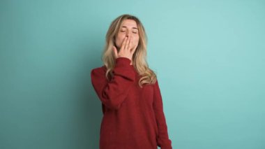 Kestane rengi kazaklı, uzun sarı saçlı mutlu genç bir kadın öpücük yolluyor ve mavi arka plandaki kameraya bakarken kalp hareketi yapıyor.