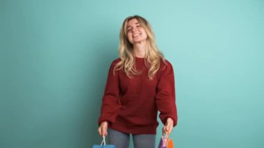 Kestane rengi kazaklı, uzun sarı saçlı gülümseyen genç kadın portresi mavi arka planda renkli kağıt alışveriş torbaları gösteriyor.