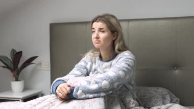 Pijamalı genç bir kadın yatağında oturur yatak odasında üzüntü ve endişe hisseder. Duygularını yansıtır.