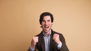 Ceketli, siyah saçlı, yumruklarını sıkan heyecanlı iş adamının portresi. Kahverengi arka planda duran kameraya bakarak başarıyı kutluyor.