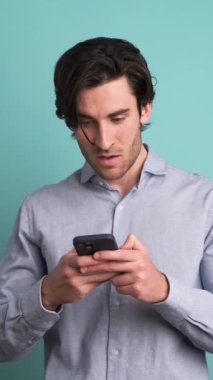 Mavi arka planda dikilirken akıllı telefondan iyi haberler aldıktan sonra şaşırmış ve kutlama yapan tişörtlü yetişkin erkek portresi