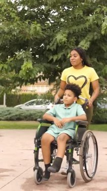 Kadın, yeşil ağaçlarla çevrili güneşli bir parkta yürürken tekerlekli sandalyedeki bir çocuğu itiyor. Dikey görüntüler