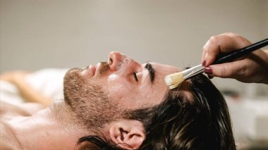 Güzellik uzmanı erkek bir müşteriye nemlendirici yüz maskesi uyguluyor, rahatlatıcı bir spa deneyimi sağlıyor.