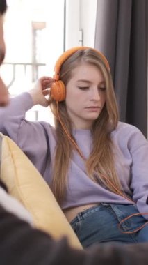Kulaklık takan genç bir kadın kanepede oturur, can sıkıntısı ve kayıtsızlığını dile getirirken müzik dinler.