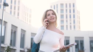 Kitap ve sırt çantasıyla kampüste güneşli bir yaz gününde yürürken akıllı telefondan konuşan neşeli genç kız öğrenci.