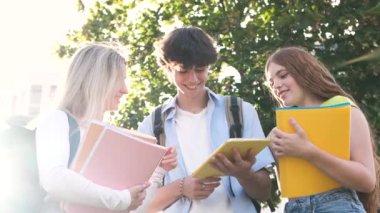 Güneşli yaz gününde üniversite kampüsünde dijital tablet kullanarak günlük giysiler içinde gülümseyen genç erkek ve bayan öğrencilerin yavaş hareket etmesi.