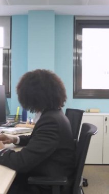 Afro saç stili olan, masa başında bilgisayar kullanan ve ortak çalışma alanındaki kameraya bakan mutlu, genç, Afrikalı, yaratıcı iş adamı.