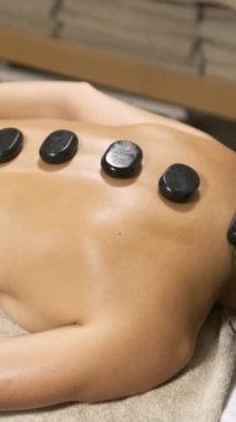 Masöz, bir masaj masasında sakin bir spa ortamında rahatlarken bir kadının sırtına sıcak taşlar yerleştiriyor. Dikey görüntüler