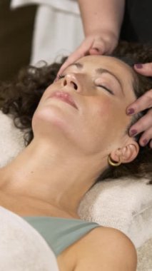 Sakin bir kaplıca ortamındaki yetenekli bir güzellik uzmanının rahatlatıcı yüz masajından zevk alan bir kadın gibi rahatlama havayı dolduruyor. Dikey görüntüler
