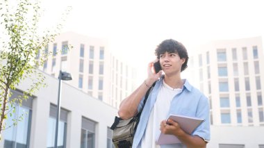 Kitap ve sırt çantasıyla kendinden emin genç bir erkek öğrenci güneşli yaz gününde üniversite binasına karşı akıllı telefondan konuşuyor.