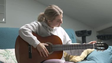 Genç bir kadın akustik gitarını akort ederken koltukta rahat bir şekilde oturur ve huzurlu bir müzik çalışmasının tadını çıkarır.