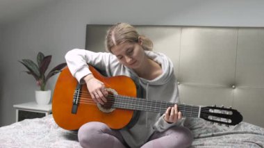 Genç bir kadın yatakta oturuyor, akustik gitar çalıyor ve rahat bir ortamda müzik becerilerini geliştiriyor.