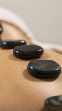 Müşteri masaj masasında rahatlatıcı bir tedaviden zevk alırken, masözün müşterilerine sıcak taşlar yerleştirmesi. Dikey görüntüler