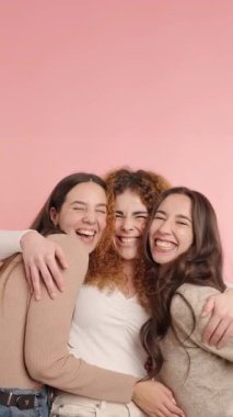 Üç neşeli bayan arkadaş kucaklaşıyor ve gülüyor, canlı pembe bir arkaplana karşı gerçek neşe ve dostluk ifade ediyorlar.