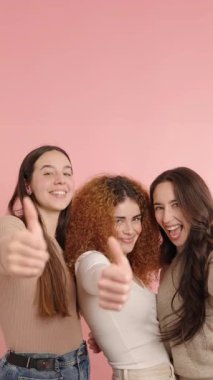 Bir grup neşeli bayan arkadaş pembe arka planda birlikte poz vererek onaylama ve onaylama jesti yapıyorlar.
