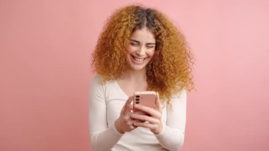 Akıllı telefonuyla kıvırcık saçlı, beyaz gömlek giyen ve canlı pembe stüdyo geçmişine gülümseyen neşeli bir milenyum kadını.