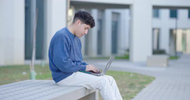 Üniversite öğrencisi kampüs bankında otururken dizüstü bilgisayara odaklanıyor, dijital öğrenme ve modern akademik yaşam tarzını somutlaştırıyor