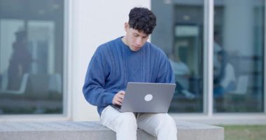 Laptop bilgisayarında çalışan, üniversite binasının önündeki bankta oturan Latin kökenli bir öğrenci.