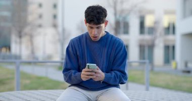 Hevesli genç adam zafer anını kutluyor, akıllı telefondan olumlu haberler alıyor, dışarıda oturuyor, zafer dolu yumruklarıyla sevinçlerini ifade ediyor.