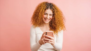 Kıvırcık saçlı, akıllı telefon kullanan, gülen pembe arka planda izole edilmiş mutlu bir genç kadın.