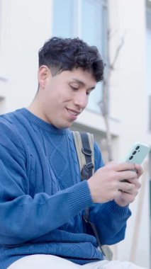 Genç Latin öğrenci akıllı telefonlara takılıyor, sosyal medyada geziniyor ve açık havada mavi süveter içinde otururken mesajlaşıyor. Dikey görüntüler