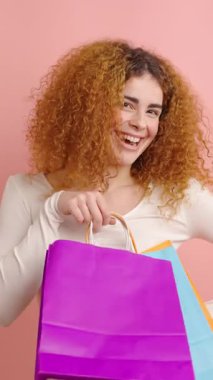 Gülümseyen kızıl saçlı kadın elinde parlak alışveriş çantaları tutarak pembe stüdyo geçmişine neşe ve heyecan katıyor perakende terapisi ve moda tarzı sergiliyor.