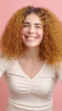 Neşeli kıvırcık saçlı kadın hediye paketini kırmızı kurdeleyle sunuyor. Yumuşak pembe arka planda duruyor. Sıcak cömertlik ve neşe ifade ediyor.