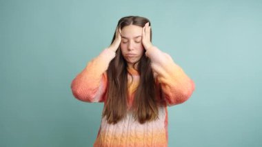 Mavi arka planda dikilirken baş ağrısından muzdarip, elleri başının üzerinde olan, sıradan giyinmiş, üzgün genç bir kadının portresi.