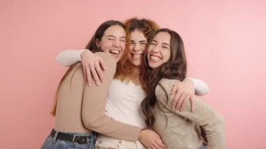 Uzun saçlı neşeli genç kadınlar pembe arka planda dikilirken kameraya sarılıyorlar.