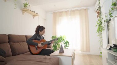 Kahverengi bir koltukta oturan kadın, ustalıkla akustik gitar çalıyor. Perdelerden süzülen güneş ışığı oturma odasının sakin ve rahat atmosferini arttırır.